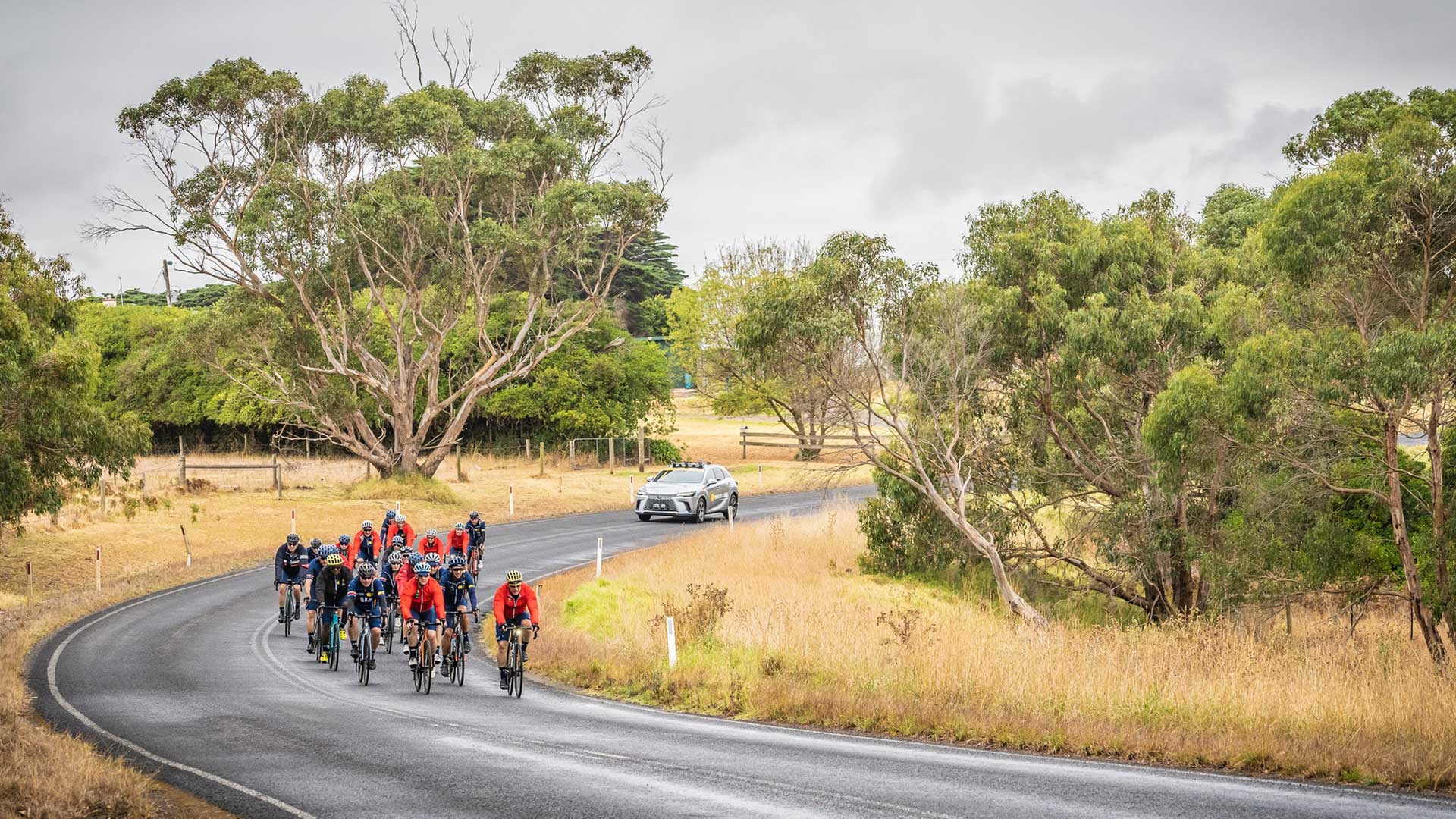 A group of cyclists ride together along a country road, with a support vehicle following behind.